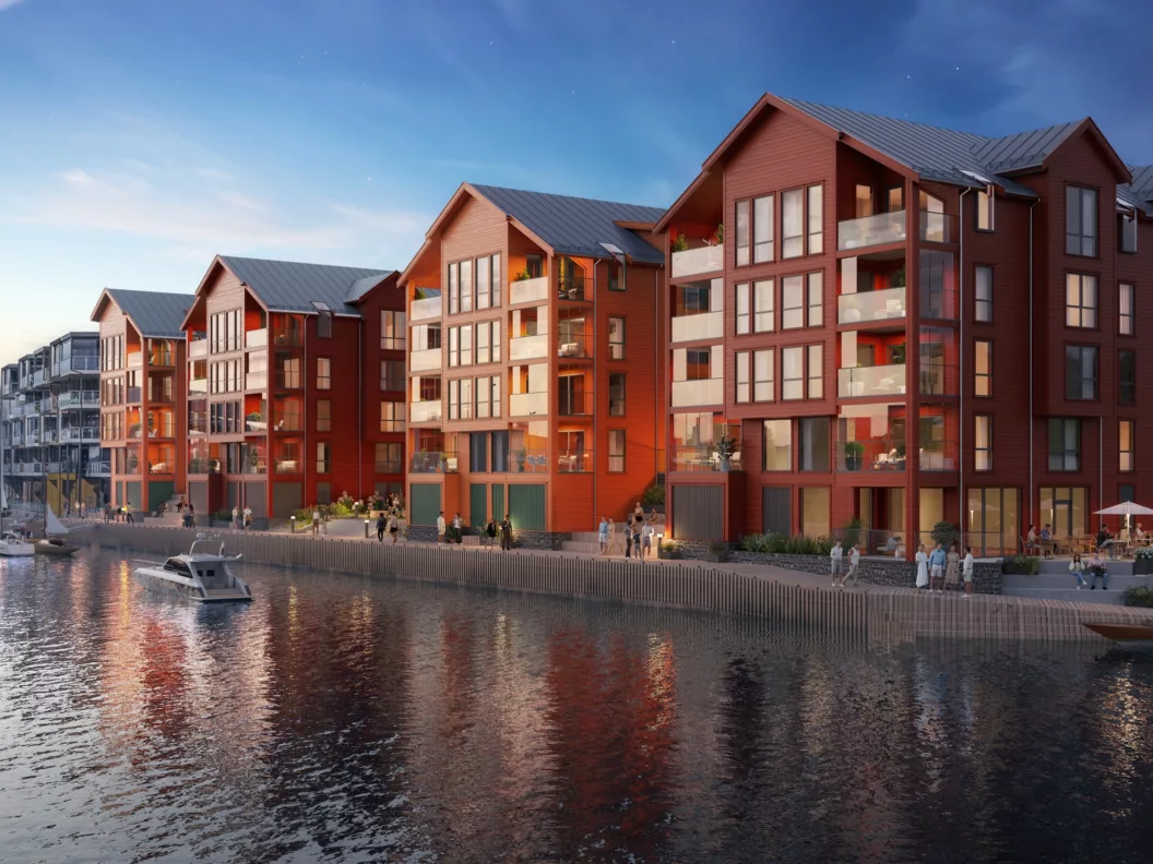 En rekke moderne røde bygninger med store vinduer langs en vannkant, med en båt som ligger til kai i nærheten og folk som går langs promenaden under en skumringshimmel.